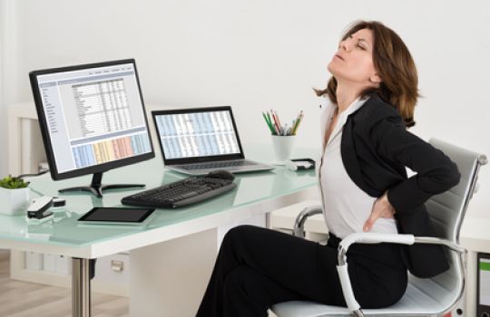Eine Frau sitzt vor ihrem Computer und fasst sich an den schmerzenden Rücken.