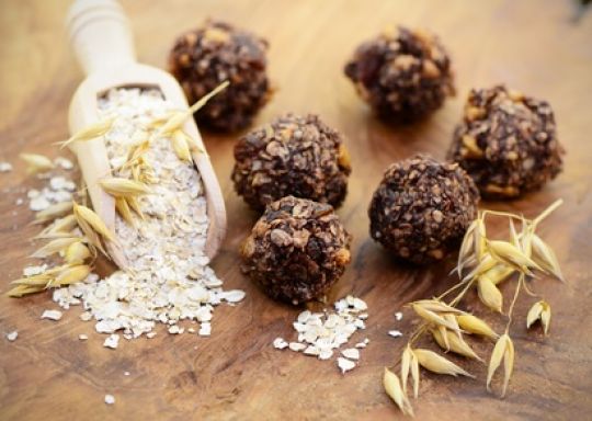 Kleine Energiebällchen liegen neben einem Holzschäufelchen mit Haferflocken.