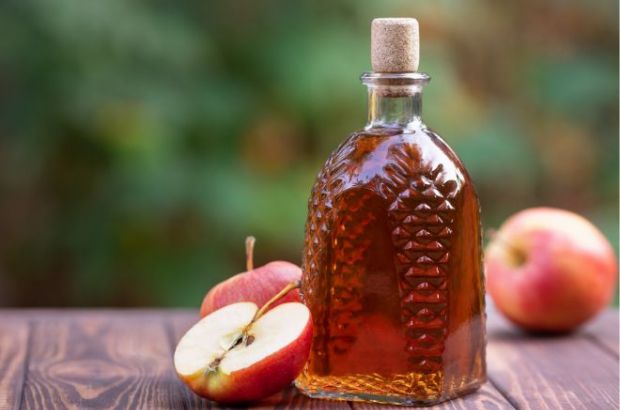 Apfelessig ist ein altes und gesundes Hausmittel. © Alter_photo Getty Images on canva Apfelessig in einer schönen Flasche steht mit Äpfeln auf einem Holztisch.